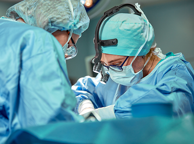 surgeons in blue scrubs using magnifiers and lights to focus on a patient being operated on