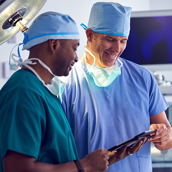 a surgeon in surgical scrubs looking at a tablet alongside a theatre assistant in a hospital office